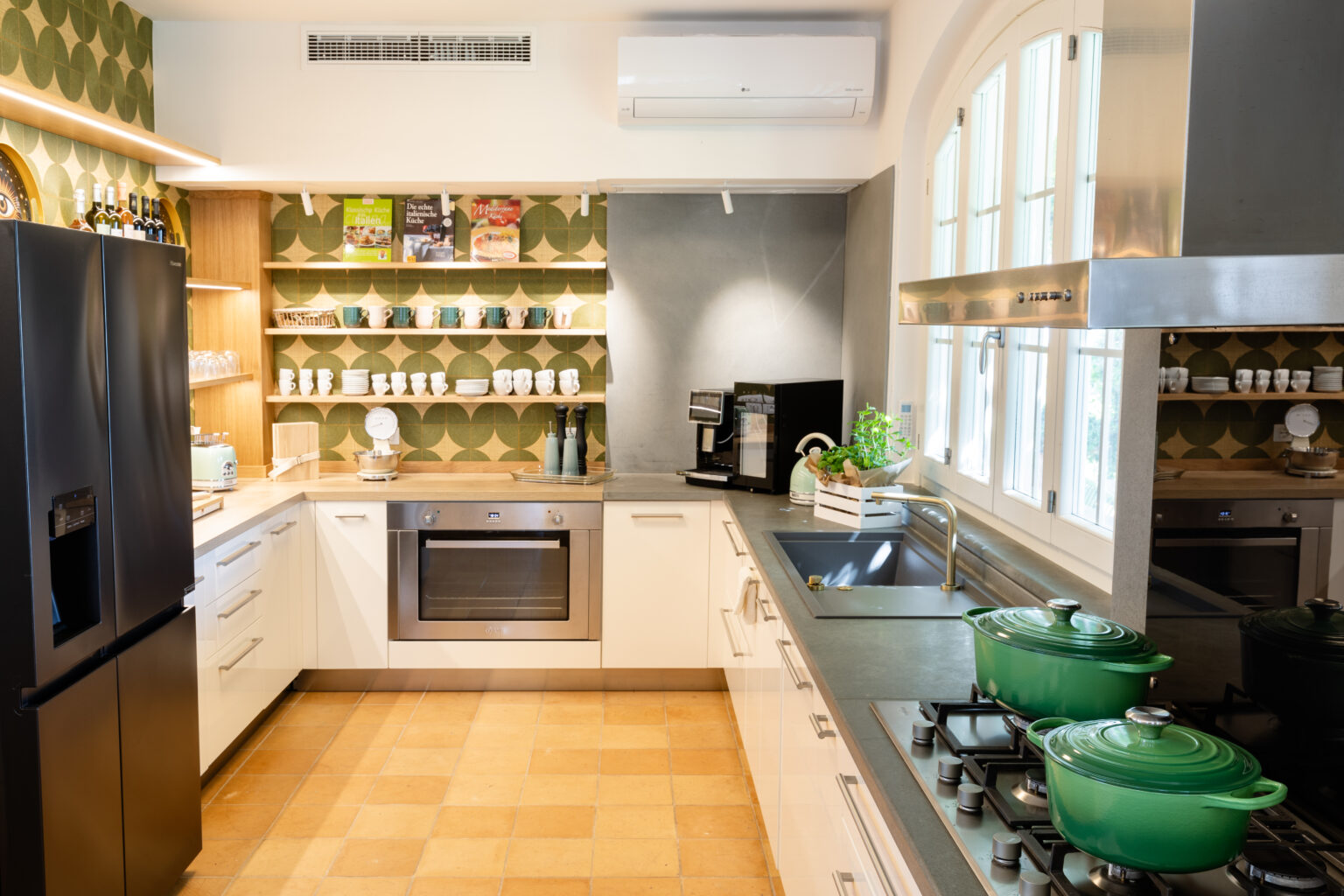 Modern kitchen interior featuring sleek cabinetry, a stylish backsplash with geometric patterns, stainless steel appliances including a refrigerator and oven, and green cookware on the stove. Natural light streams in through large windows, enhancing the inviting atmosphere. | Only on SopranoVillas