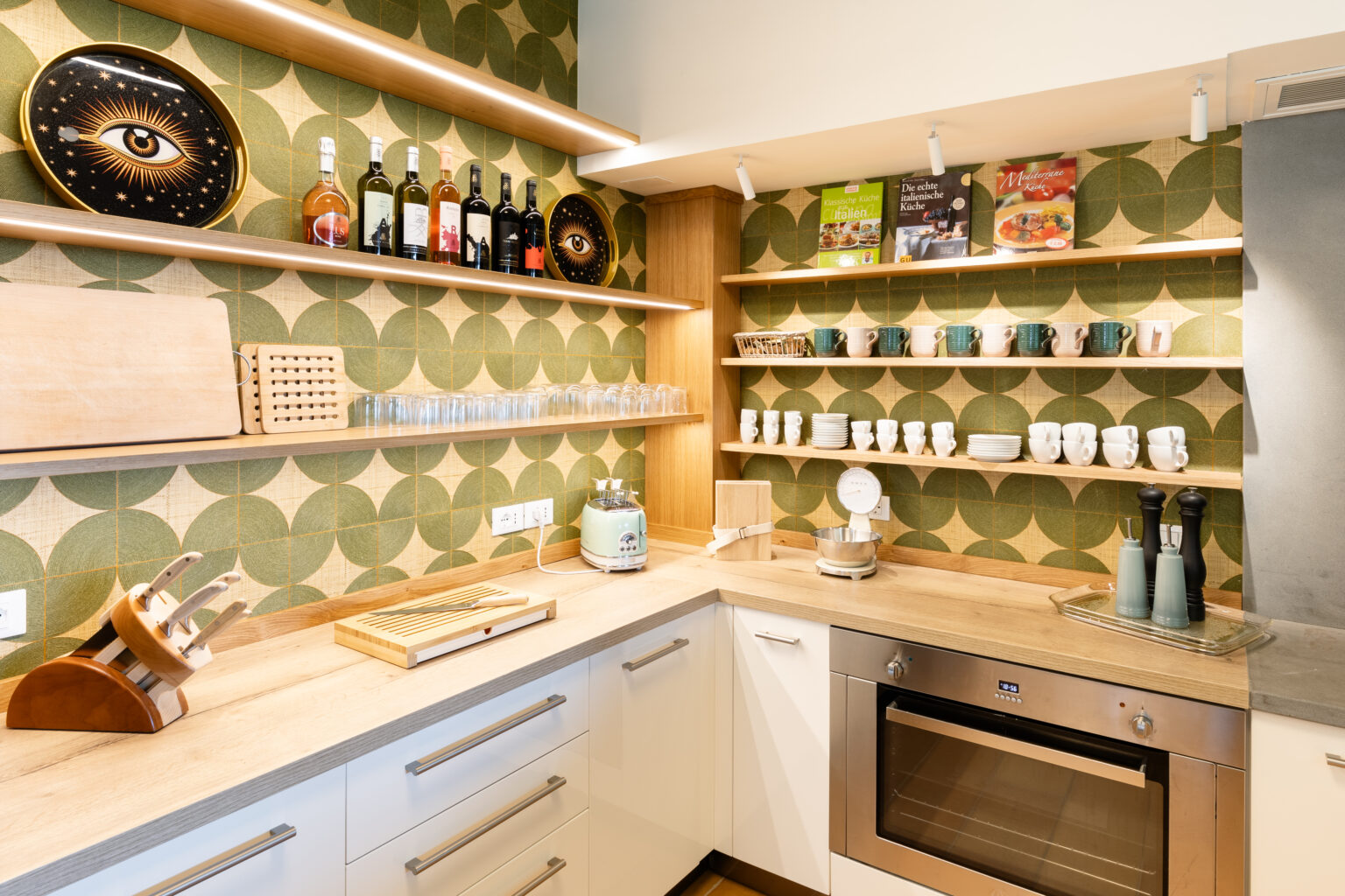 Modern kitchen interior featuring stylish shelving with wine bottles, glassware, and cookbooks, complemented by a patterned green wallpaper. The countertop displays kitchen utensils, a toaster, and a scale, creating a functional and aesthetically pleasing space. | Only on SopranoVillas