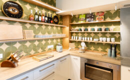 Modern kitchen interior featuring stylish shelving with wine bottles, glassware, and cookbooks, complemented by a patterned green wallpaper. The countertop displays kitchen utensils, a toaster, and a scale, creating a functional and aesthetically pleasing space.
