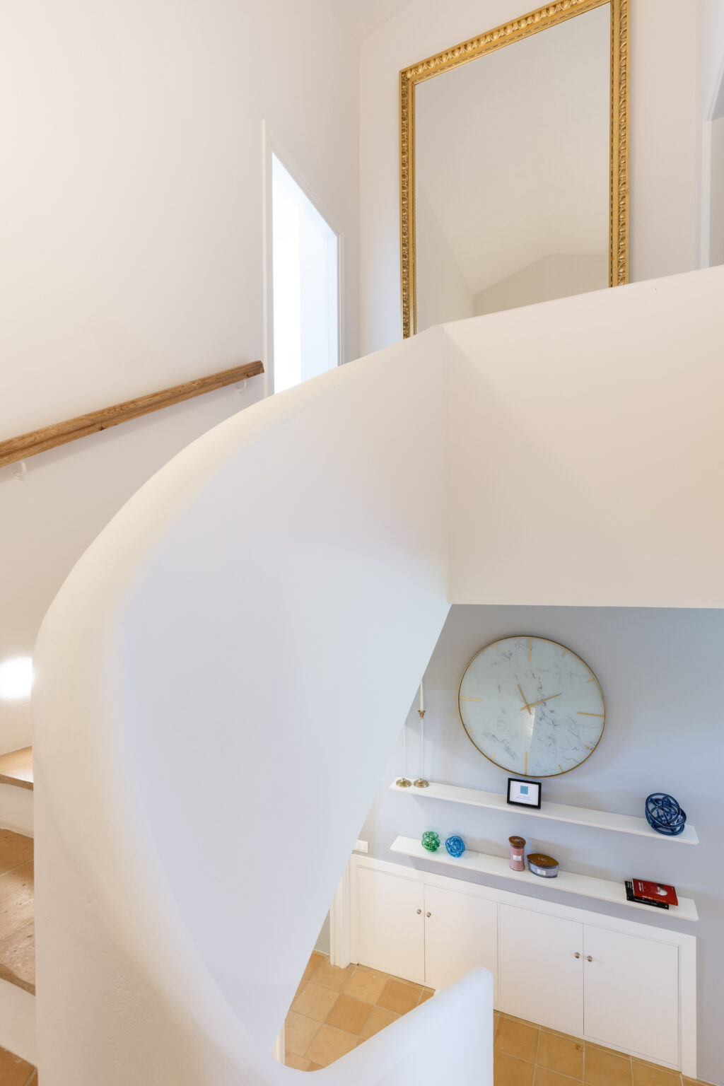 Interior view of a modern staircase with a curved white wall and wooden handrail, featuring a large gold-framed mirror above. The space includes a stylish clock on the wall, decorative items on a shelf, and a light-colored tiled floor, creating a bright and inviting atmosphere. | Only on SopranoVillas