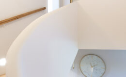 Interior view of a modern staircase with a curved white wall and wooden handrail, featuring a large gold-framed mirror above. The space includes a stylish clock on the wall, decorative items on a shelf, and a light-colored tiled floor, creating a bright and inviting atmosphere.