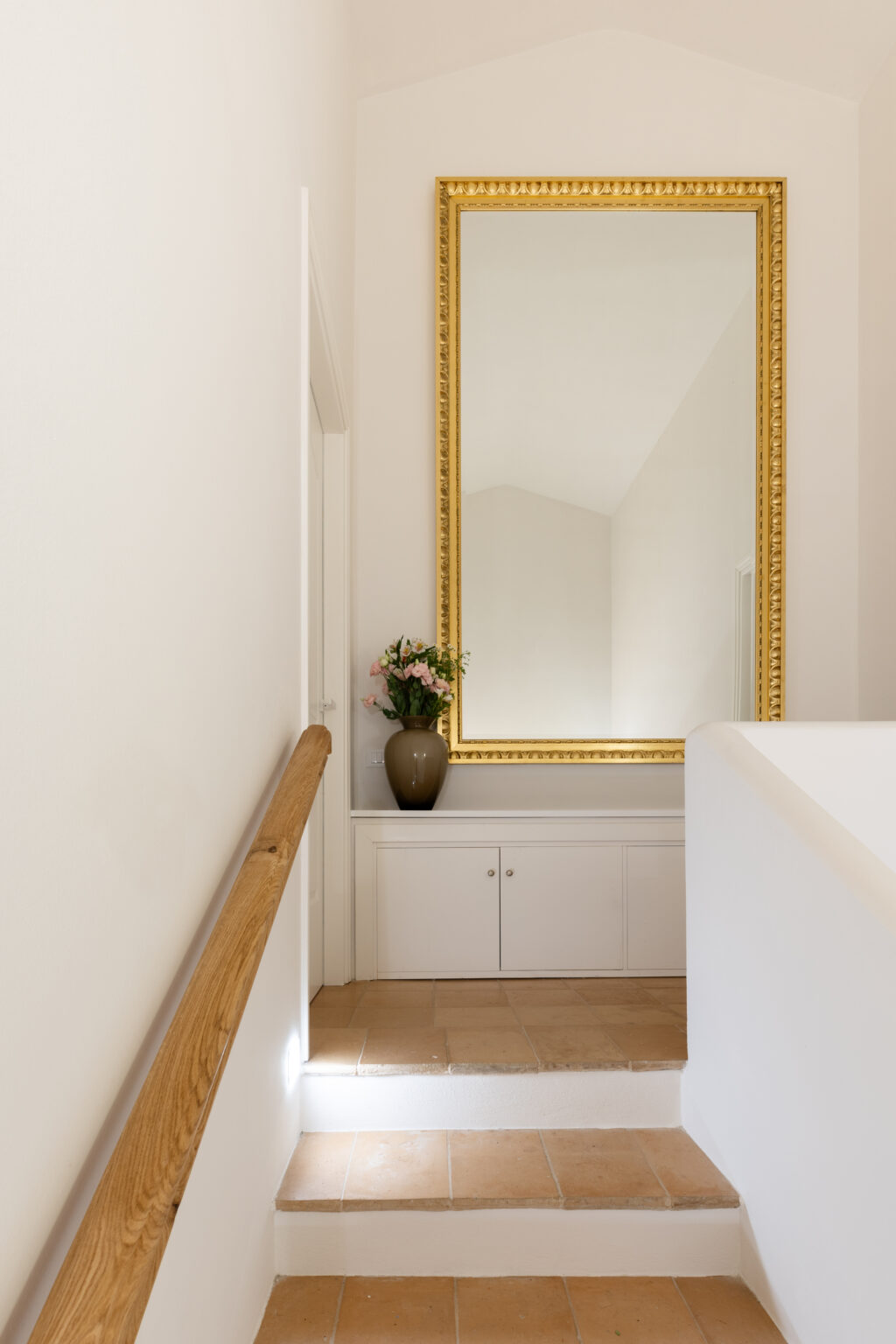 Elegant staircase leading to a well-lit hallway, featuring a large ornate gold-framed mirror and a decorative vase with flowers on a white console table. The warm-toned tile floor complements the minimalist decor and wooden handrail. | Only on SopranoVillas