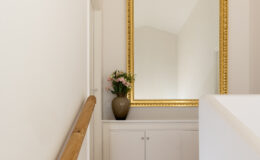 Elegant staircase leading to a well-lit hallway, featuring a large ornate gold-framed mirror and a decorative vase with flowers on a white console table. The warm-toned tile floor complements the minimalist decor and wooden handrail.
