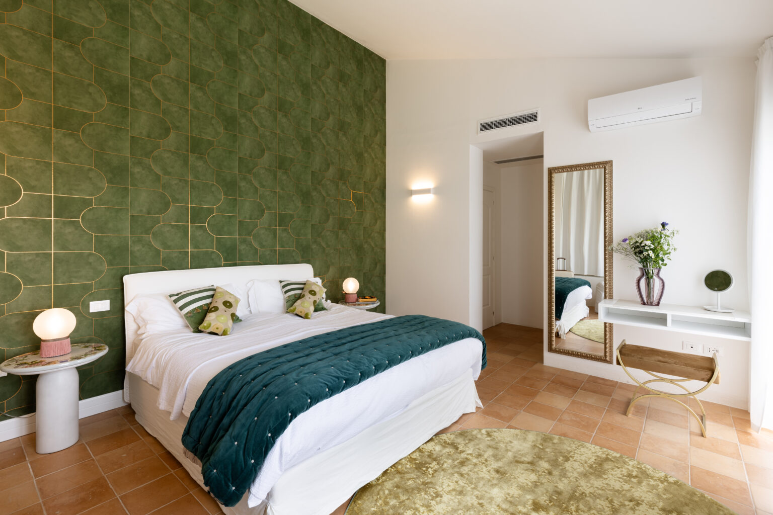 Modern bedroom featuring a green patterned accent wall, a comfortable bed with decorative pillows, and stylish furnishings including a round rug, a bedside lamp, and a mirror with a floral arrangement. The room has natural light and a contemporary design aesthetic. | Only on SopranoVillas