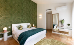 Modern bedroom featuring a green patterned accent wall, a comfortable bed with decorative pillows, and stylish furnishings including a round rug, a bedside lamp, and a mirror with a floral arrangement. The room has natural light and a contemporary design aesthetic.