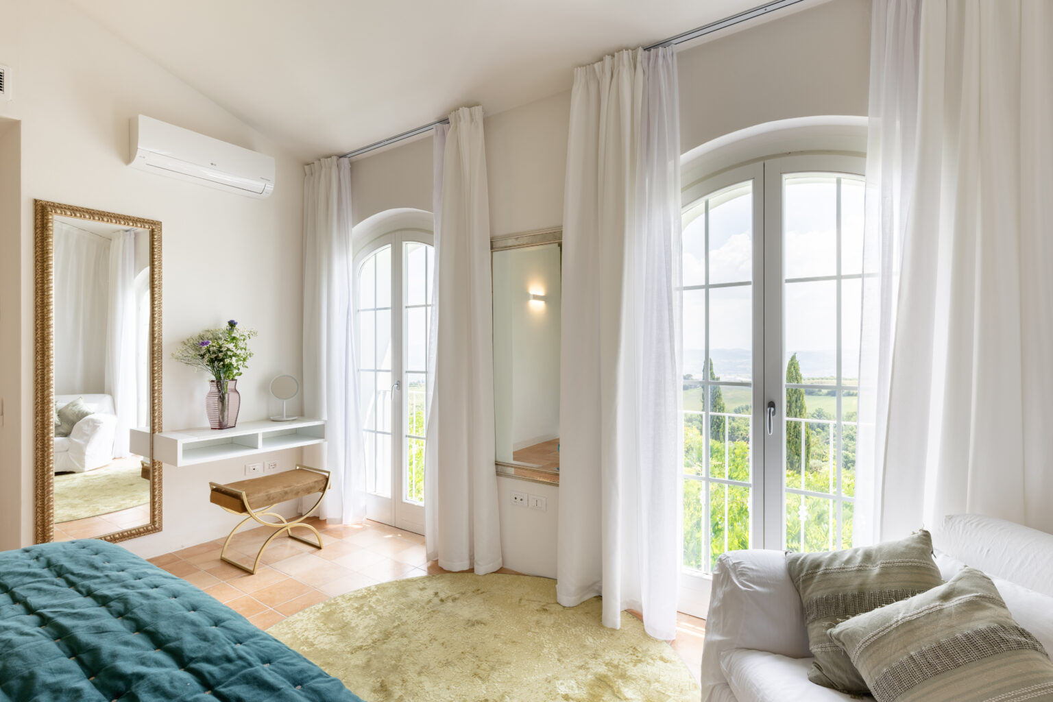 Bright and airy bedroom featuring large windows with scenic views, soft white curtains, a plush teal bedspread, and a stylish gold-accented mirror. The room includes a minimalist white vanity, a small wooden bench, and a cozy seating area with decorative pillows, creating a serene and inviting atmosphere. | Only on SopranoVillas