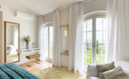 Bright and airy bedroom featuring large windows with scenic views, soft white curtains, a plush teal bedspread, and a stylish gold-accented mirror. The room includes a minimalist white vanity, a small wooden bench, and a cozy seating area with decorative pillows, creating a serene and inviting atmosphere.