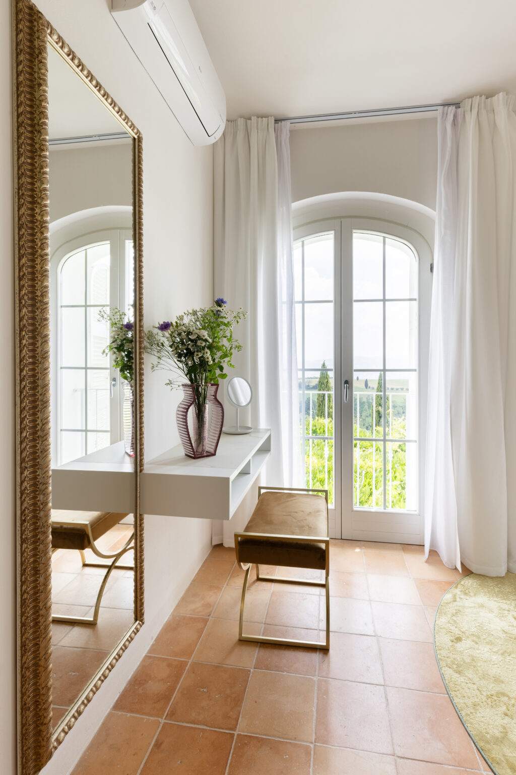Bright and airy bedroom corner featuring a stylish vanity table with a mirror, a decorative vase of flowers, and a plush upholstered stool. Large windows with white curtains allow natural light to fill the space, complemented by terracotta tiled flooring and a soft round rug. | Only on SopranoVillas