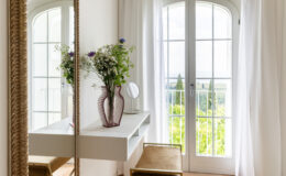 Bright and airy bedroom corner featuring a stylish vanity table with a mirror, a decorative vase of flowers, and a plush upholstered stool. Large windows with white curtains allow natural light to fill the space, complemented by terracotta tiled flooring and a soft round rug.