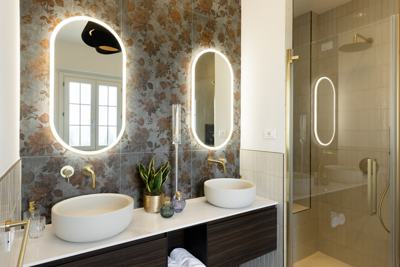 Modern bathroom featuring dual oval mirrors with integrated lighting, elegant gold fixtures, and stylish vessel sinks. The wall is adorned with floral-patterned tiles, complemented by a glass shower enclosure and a small indoor plant for a touch of greenery. | Only on SopranoVillas