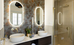 Modern bathroom featuring dual oval mirrors with integrated lighting, elegant gold fixtures, and stylish vessel sinks. The wall is adorned with floral-patterned tiles, complemented by a glass shower enclosure and a small indoor plant for a touch of greenery.