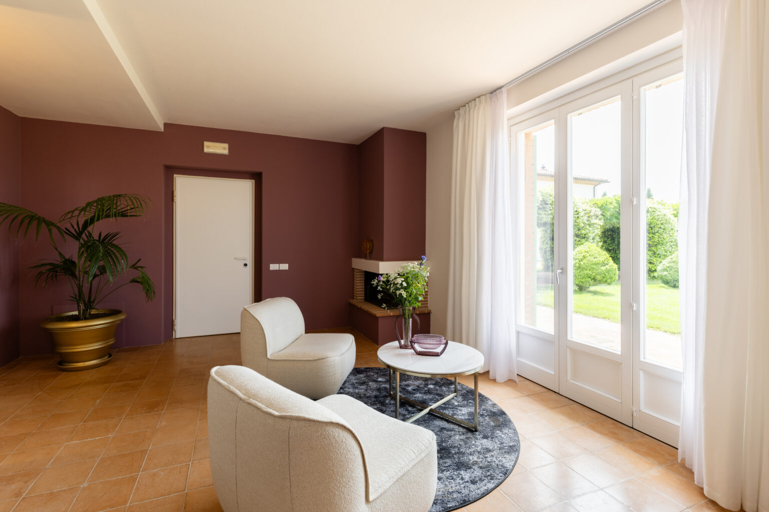 Modern living room with beige armchairs, a round coffee table, and a potted plant, featuring a large window with sheer curtains that opens to a green garden. The walls are painted in a warm mauve color, creating a cozy atmosphere. | Only on SopranoVillas