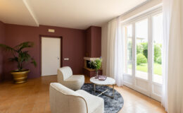 Modern living room with beige armchairs, a round coffee table, and a potted plant, featuring a large window with sheer curtains that opens to a green garden. The walls are painted in a warm mauve color, creating a cozy atmosphere.