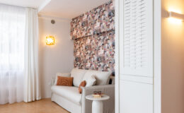 Modern living room featuring a light-colored sofa with decorative pillows, a stylish round rug, and a minimalist side table with books and glasses. The wall is adorned with a colorful patterned wallpaper, complemented by sheer curtains and a wall-mounted light fixture, creating a cozy and inviting atmosphere.