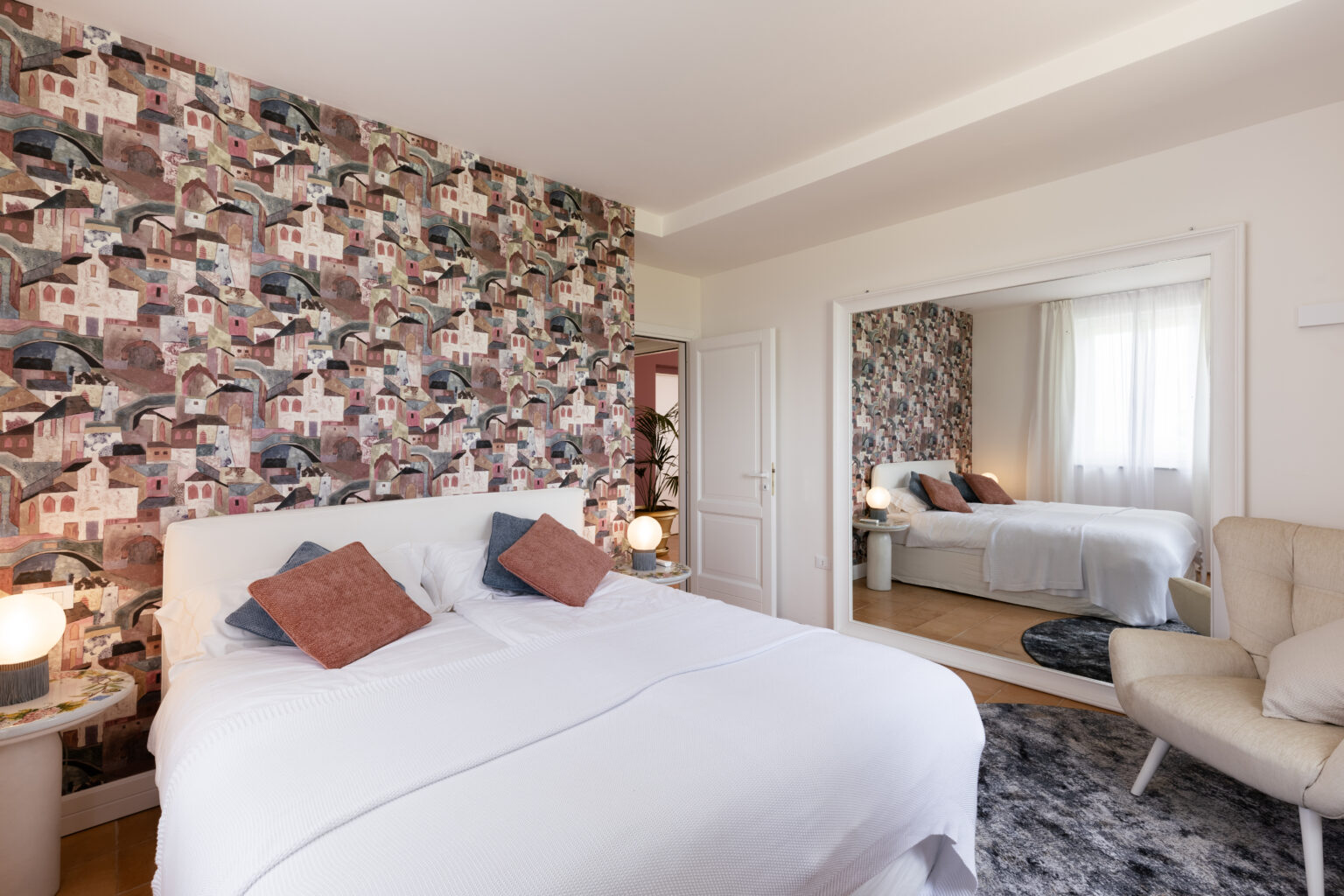 Modern bedroom featuring a stylish design with a patterned accent wall, a plush white bed adorned with decorative pillows, and a cozy reading chair. The room includes a large mirror and soft lighting, creating a warm and inviting atmosphere. | Only on SopranoVillas