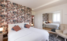 Modern bedroom featuring a stylish design with a patterned accent wall, a plush white bed adorned with decorative pillows, and a cozy reading chair. The room includes a large mirror and soft lighting, creating a warm and inviting atmosphere.