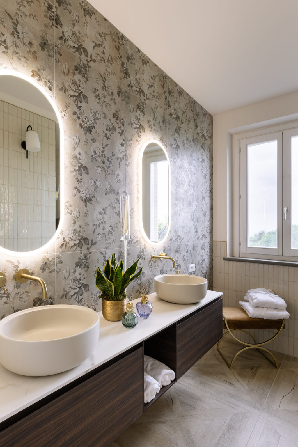 Modern bathroom interior featuring dual oval sinks, elegant gold faucets, and a floral wallpaper accent. A potted plant adds a touch of greenery, while decorative bottles and neatly stacked towels enhance the aesthetic. Natural light streams in through the window, creating a bright and inviting atmosphere. | Only on SopranoVillas