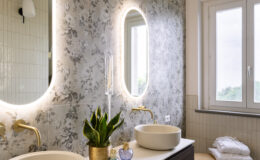 Modern bathroom interior featuring dual oval sinks, elegant gold faucets, and a floral wallpaper accent. A potted plant adds a touch of greenery, while decorative bottles and neatly stacked towels enhance the aesthetic. Natural light streams in through the window, creating a bright and inviting atmosphere.