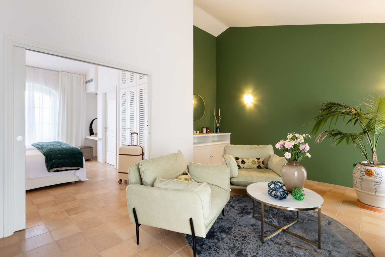 Cozy living area featuring two light green sofas, a round coffee table with decorative items, and a potted plant. The space is connected to a bedroom with a single bed and suitcase visible through an open doorway. A green accent wall adds a modern touch, complemented by soft lighting and neutral-colored flooring. | Only on SopranoVillas