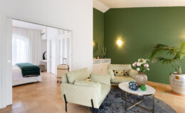 Cozy living area featuring two light green sofas, a round coffee table with decorative items, and a potted plant. The space is connected to a bedroom with a single bed and suitcase visible through an open doorway. A green accent wall adds a modern touch, complemented by soft lighting and neutral-colored flooring.