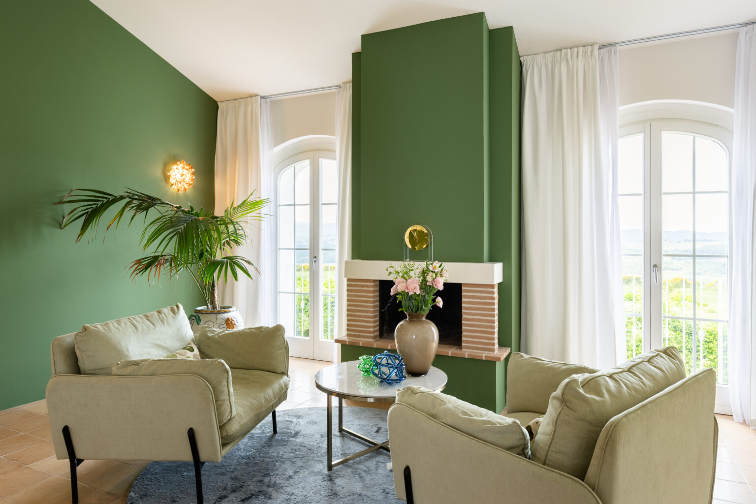Cozy living room featuring a green accent wall, soft beige sofas, a round coffee table, and a decorative vase with flowers. Natural light floods in from large windows, enhancing the serene atmosphere complemented by indoor plants and stylish decor elements. | Only on SopranoVillas
