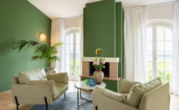 Cozy living room featuring a green accent wall, soft beige sofas, a round coffee table, and a decorative vase with flowers. Natural light floods in from large windows, enhancing the serene atmosphere complemented by indoor plants and stylish decor elements.
