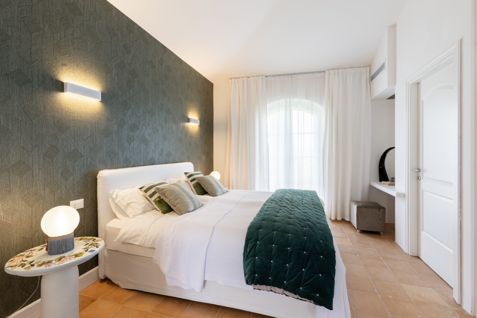 Modern bedroom interior featuring a white bed with green decorative pillows and a textured green accent wall, illuminated by stylish wall sconces. A round side table with a floral design and a soft light fixture complements the decor, while sheer curtains allow natural light to fill the room. | Only on SopranoVillas