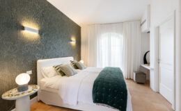 Modern bedroom interior featuring a white bed with green decorative pillows and a textured green accent wall, illuminated by stylish wall sconces. A round side table with a floral design and a soft light fixture complements the decor, while sheer curtains allow natural light to fill the room.