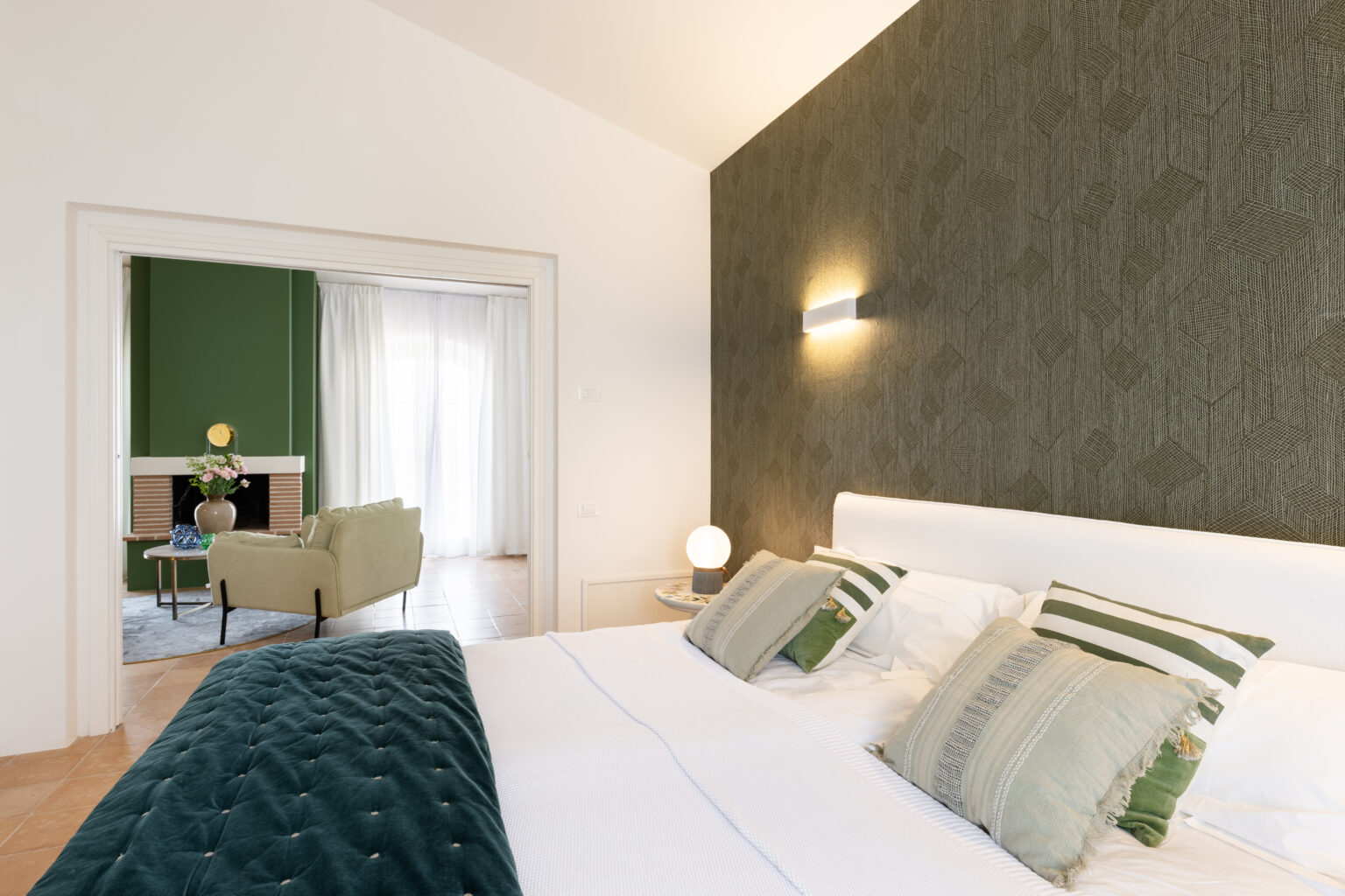 Modern bedroom interior featuring a plush green bedspread, decorative pillows, and a textured dark green accent wall. A cozy living area is visible in the background, showcasing a green sofa, a decorative table, and natural light filtering through sheer curtains. | Only on SopranoVillas