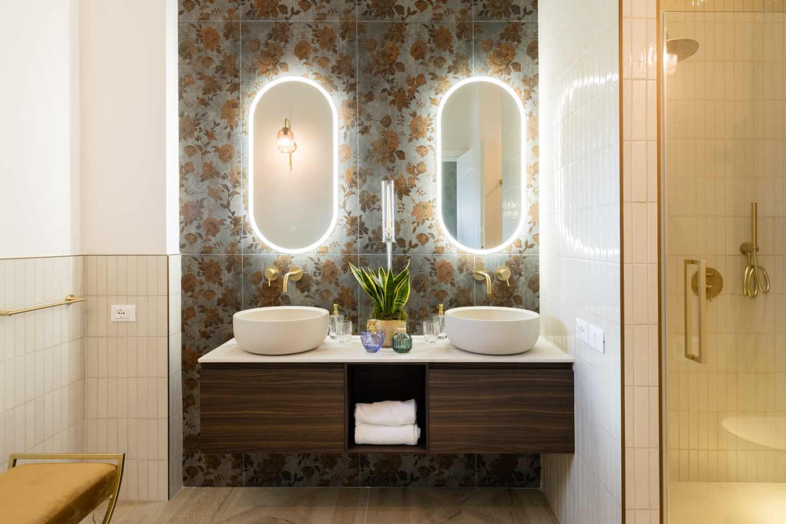Modern bathroom design featuring dual vessel sinks, elegant gold fixtures, and a floral accent wall. The space includes backlit mirrors, decorative plants, and a stylish wooden vanity with storage. A glass shower enclosure complements the contemporary aesthetic. | Only on SopranoVillas