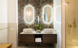 Modern bathroom design featuring dual vessel sinks, elegant gold fixtures, and a floral accent wall. The space includes backlit mirrors, decorative plants, and a stylish wooden vanity with storage. A glass shower enclosure complements the contemporary aesthetic.