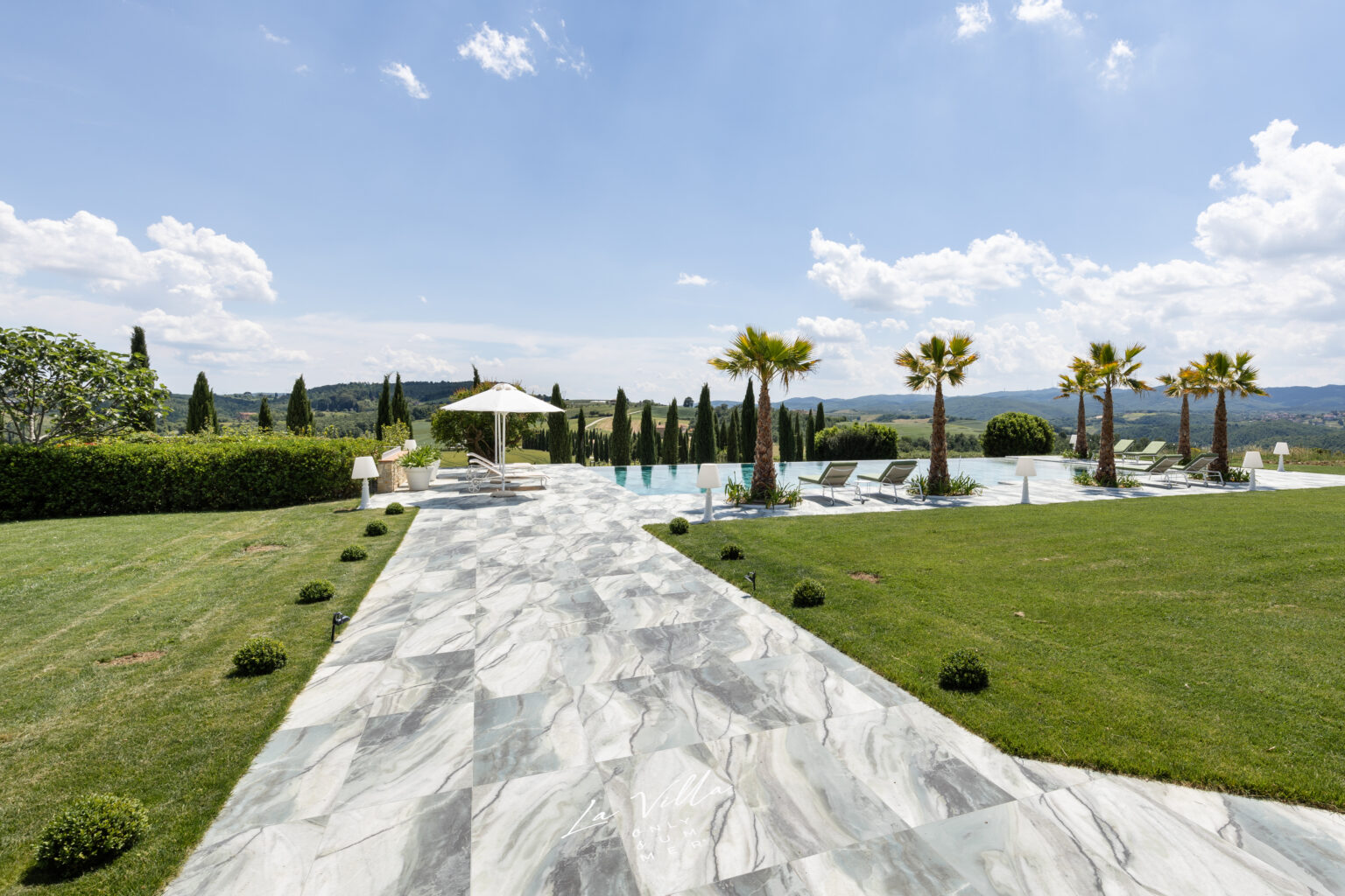 Luxurious outdoor pool area featuring a marble pathway, lush green lawn, palm trees, and sun loungers, with scenic hills and a blue sky in the background, perfect for relaxation and leisure. | Only on SopranoVillas