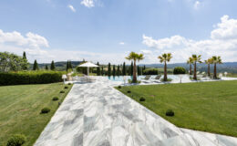 Luxurious outdoor pool area featuring a marble pathway, lush green lawn, palm trees, and sun loungers, with scenic hills and a blue sky in the background, perfect for relaxation and leisure.