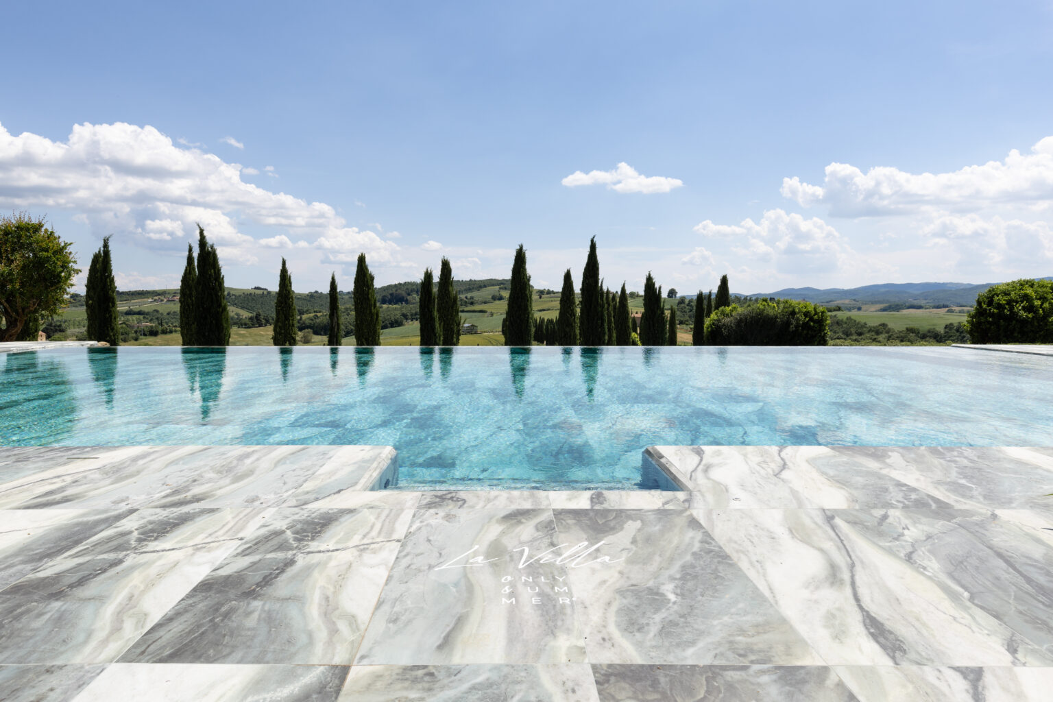 Luxurious infinity pool with clear turquoise water, surrounded by cypress trees and rolling hills under a bright blue sky. Elegant marble pool deck enhances the serene outdoor setting, perfect for relaxation and leisure. | Only on SopranoVillas