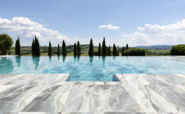 Luxurious infinity pool with clear turquoise water, surrounded by cypress trees and rolling hills under a bright blue sky. Elegant marble pool deck enhances the serene outdoor setting, perfect for relaxation and leisure.