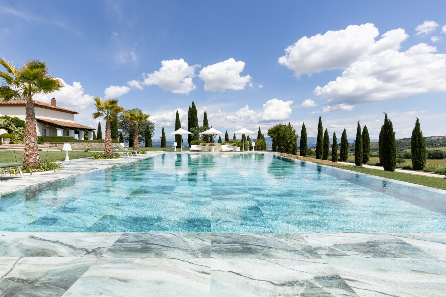 Luxurious infinity pool surrounded by palm trees and cypress trees, with a scenic view of the countryside and a clear blue sky, perfect for relaxation and outdoor living. | Only on SopranoVillas