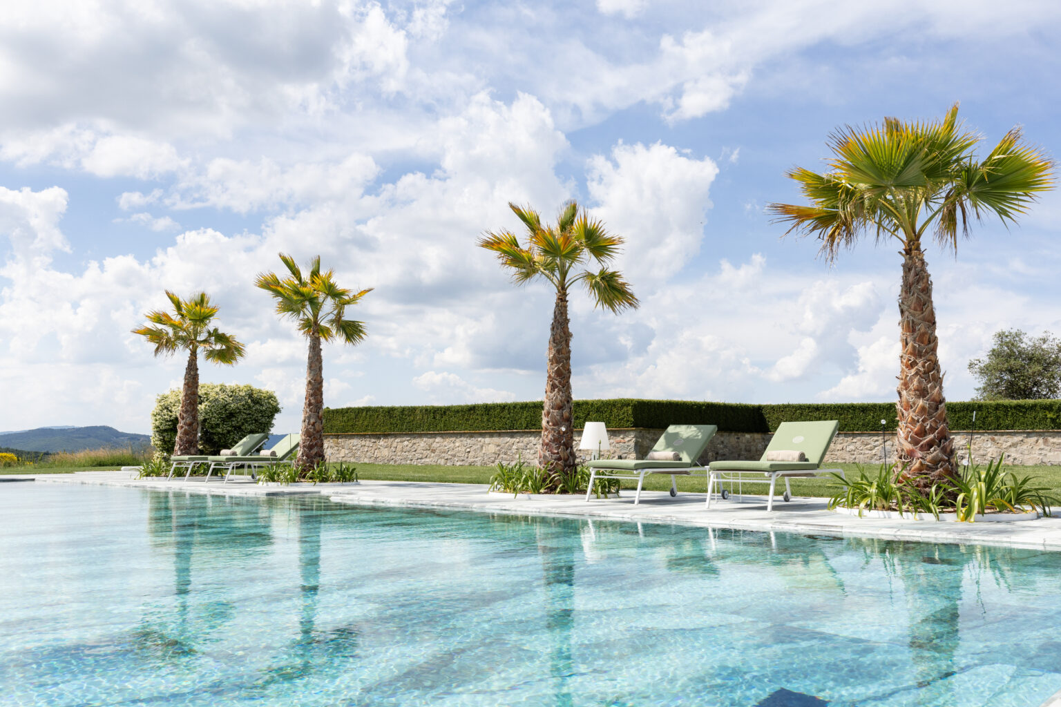 Serene outdoor swimming pool surrounded by palm trees and sun loungers, under a bright blue sky with fluffy clouds, showcasing a luxurious relaxation area in a scenic landscape. | Only on SopranoVillas