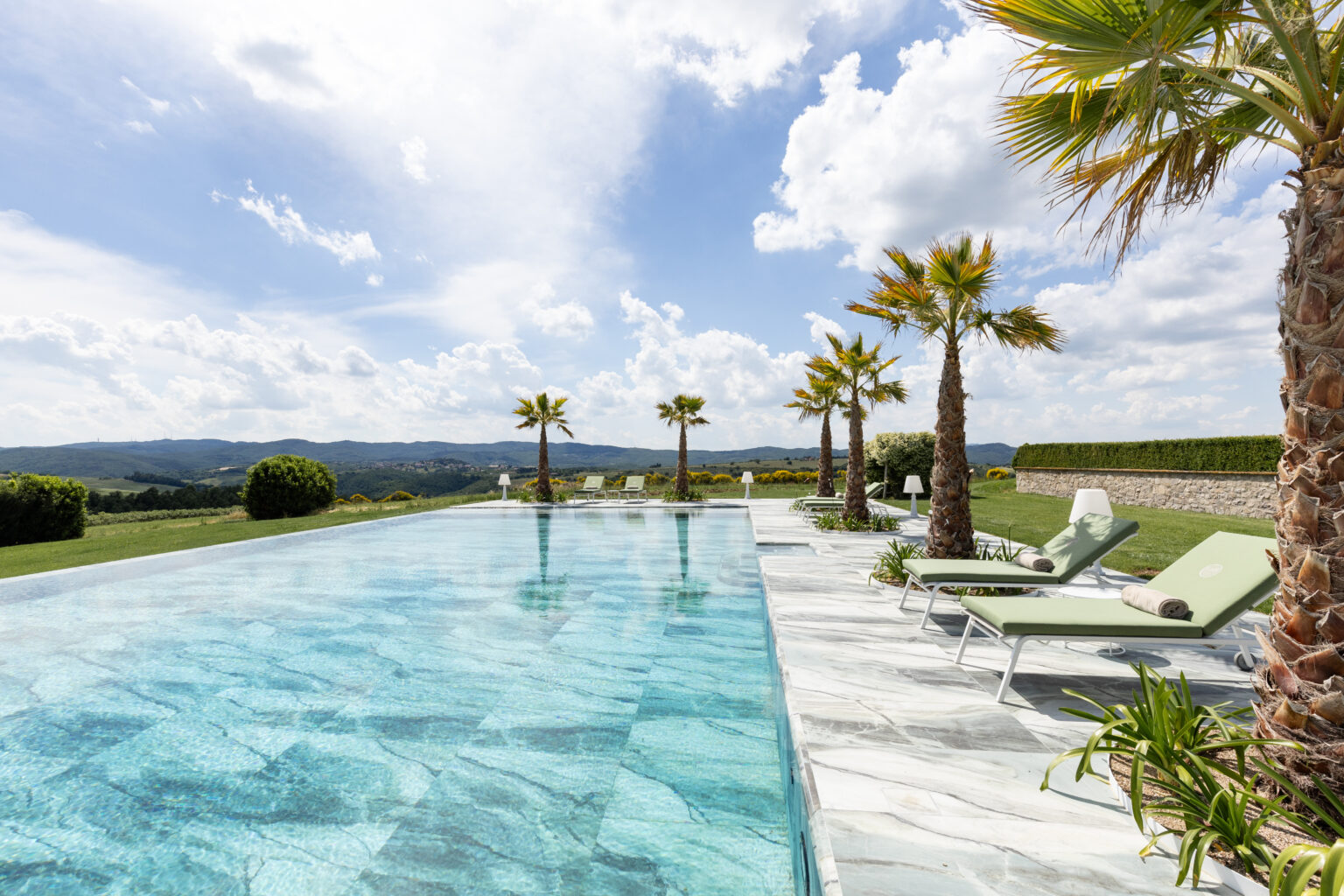 Luxurious infinity pool with palm trees, overlooking rolling hills under a blue sky with clouds. Sun loungers are placed beside the pool, creating a tranquil outdoor relaxation space. Perfect for vacation or resort imagery. | Only on SopranoVillas