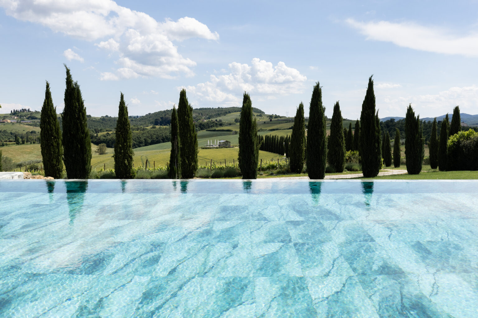 Scenic view of a tranquil infinity pool reflecting clear blue water, surrounded by tall cypress trees and rolling green hills under a bright sky in Tuscany, Italy. Ideal for luxury travel and relaxation imagery. | Only on SopranoVillas