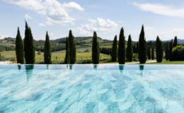 Scenic view of a tranquil infinity pool reflecting clear blue water, surrounded by tall cypress trees and rolling green hills under a bright sky in Tuscany, Italy. Ideal for luxury travel and relaxation imagery.