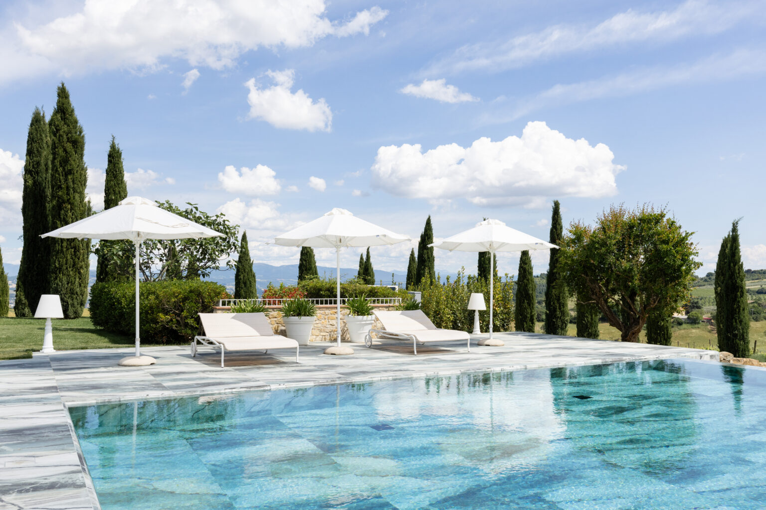 Luxury outdoor pool area with sun loungers and white umbrellas, surrounded by lush greenery and tall cypress trees under a bright blue sky with fluffy clouds. Ideal for relaxation and leisure. | Only on SopranoVillas