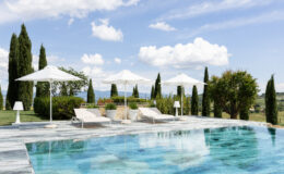 Luxury outdoor pool area with sun loungers and white umbrellas, surrounded by lush greenery and tall cypress trees under a bright blue sky with fluffy clouds. Ideal for relaxation and leisure.