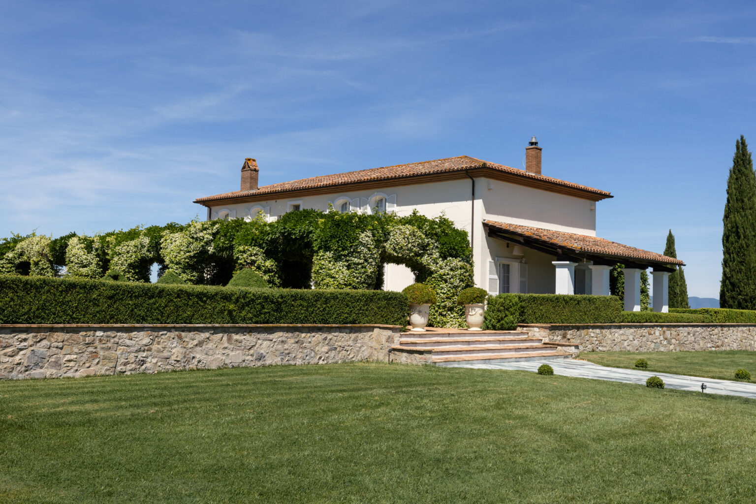 Beautiful Italian villa surrounded by lush greenery and manicured hedges, featuring a stone wall and elegant steps leading to a terrace, under a clear blue sky. Ideal for luxury real estate or vacation rental listings. | Only on SopranoVillas