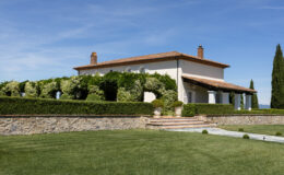 Beautiful Italian villa surrounded by lush greenery and manicured hedges, featuring a stone wall and elegant steps leading to a terrace, under a clear blue sky. Ideal for luxury real estate or vacation rental listings.