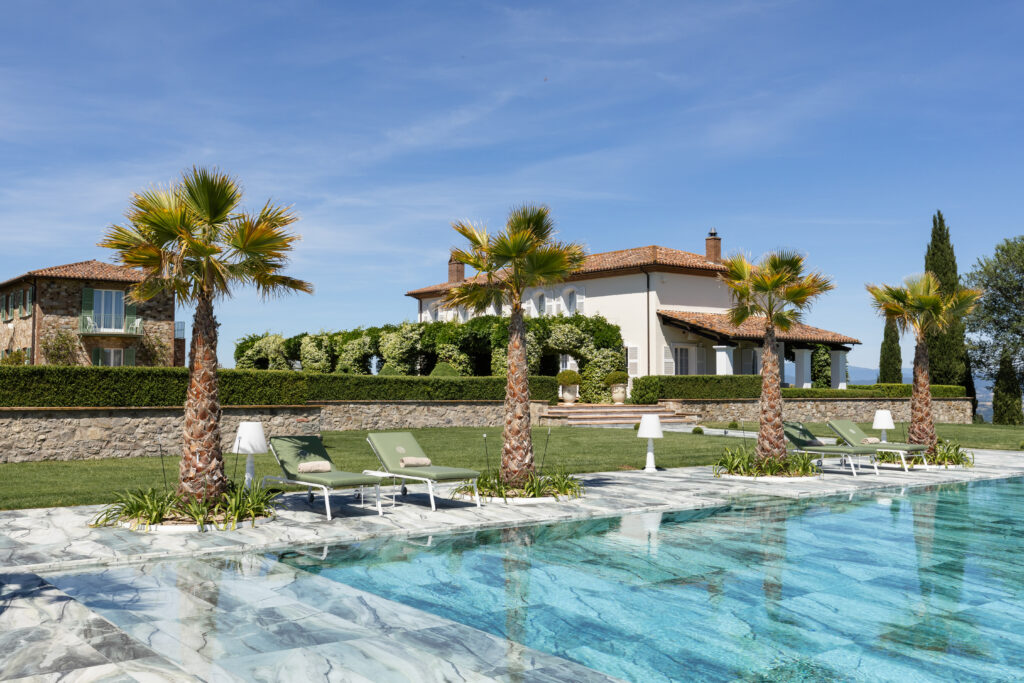 Luxury villa with a swimming pool surrounded by palm trees and sun loungers, set in a picturesque landscape under a clear blue sky. The elegant architecture features a stone facade and lush greenery, creating a serene outdoor retreat. | Only on SopranoVillas