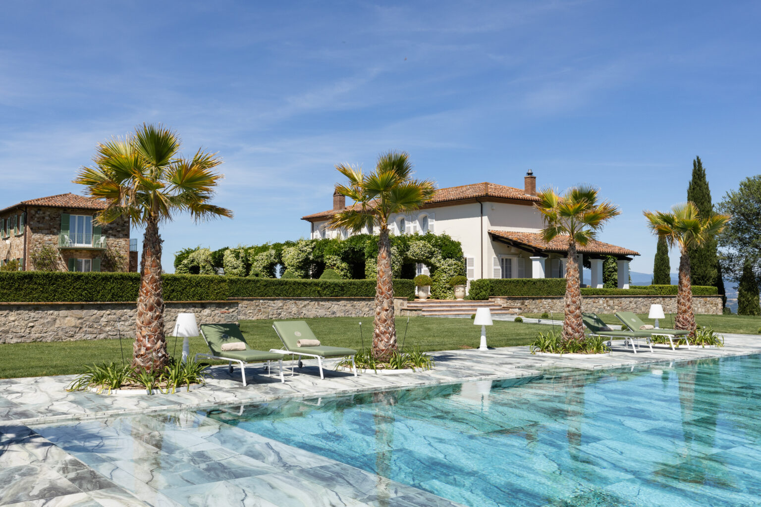 Luxury villa with a swimming pool surrounded by palm trees and sun loungers, set in a picturesque landscape under a clear blue sky. The elegant architecture features a stone facade and lush greenery, creating a serene outdoor retreat. | Only on SopranoVillas