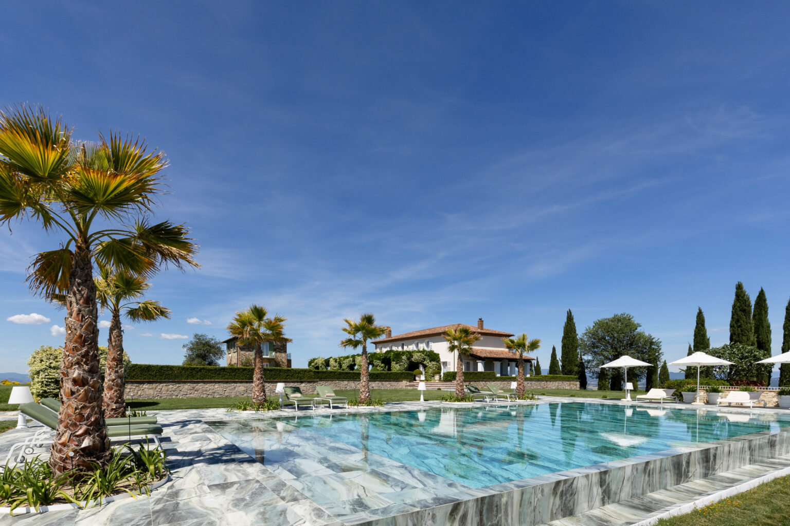 Luxurious outdoor pool surrounded by palm trees and lounge chairs, with a scenic villa and blue sky in the background, perfect for a relaxing getaway. | Only on SopranoVillas