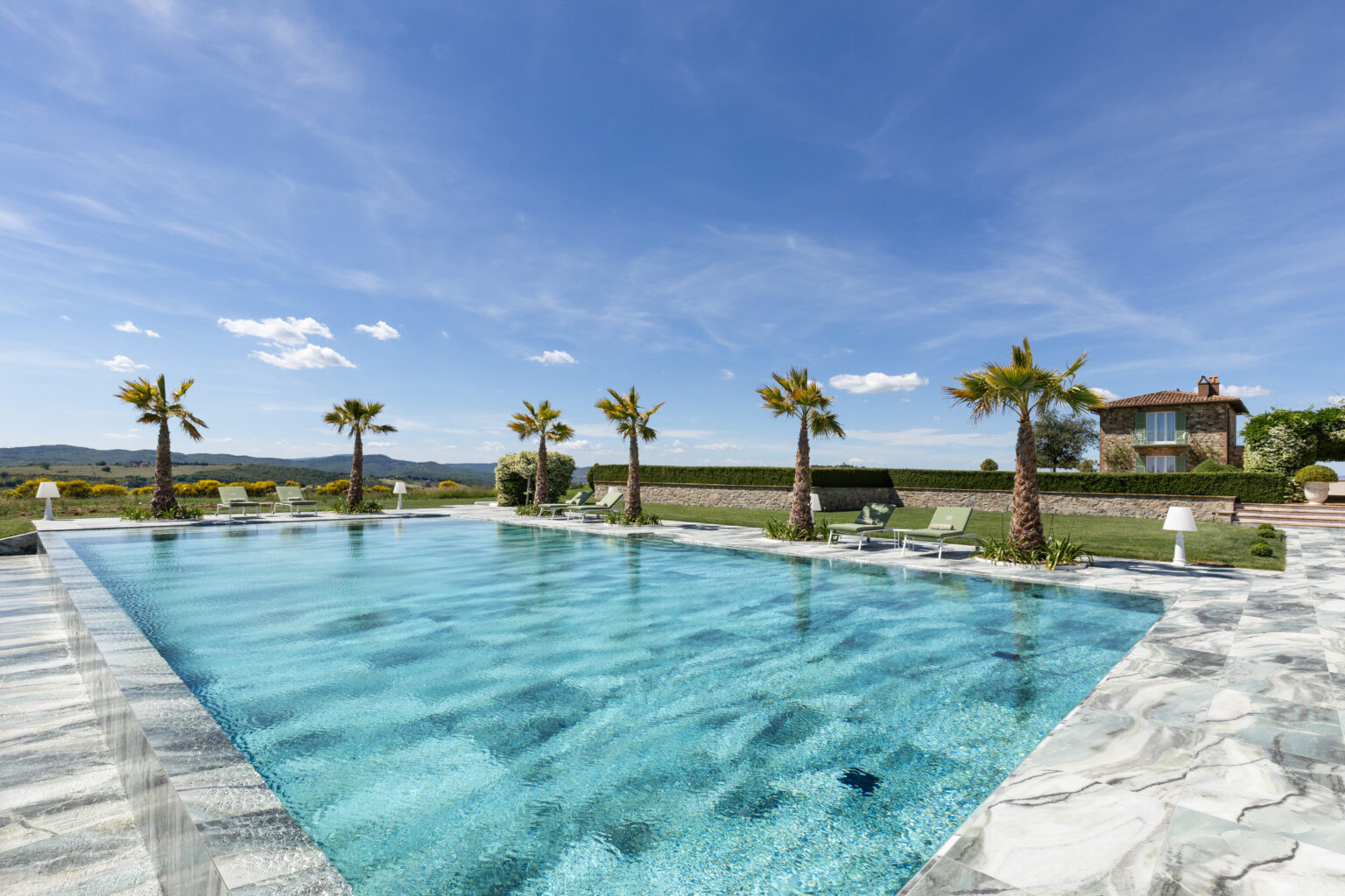 Stunning outdoor swimming pool surrounded by palm trees, with clear blue water reflecting the sky, set against a picturesque landscape and a charming stone house in the background. Ideal for luxury vacation rentals or serene retreats. | Only on SopranoVillas