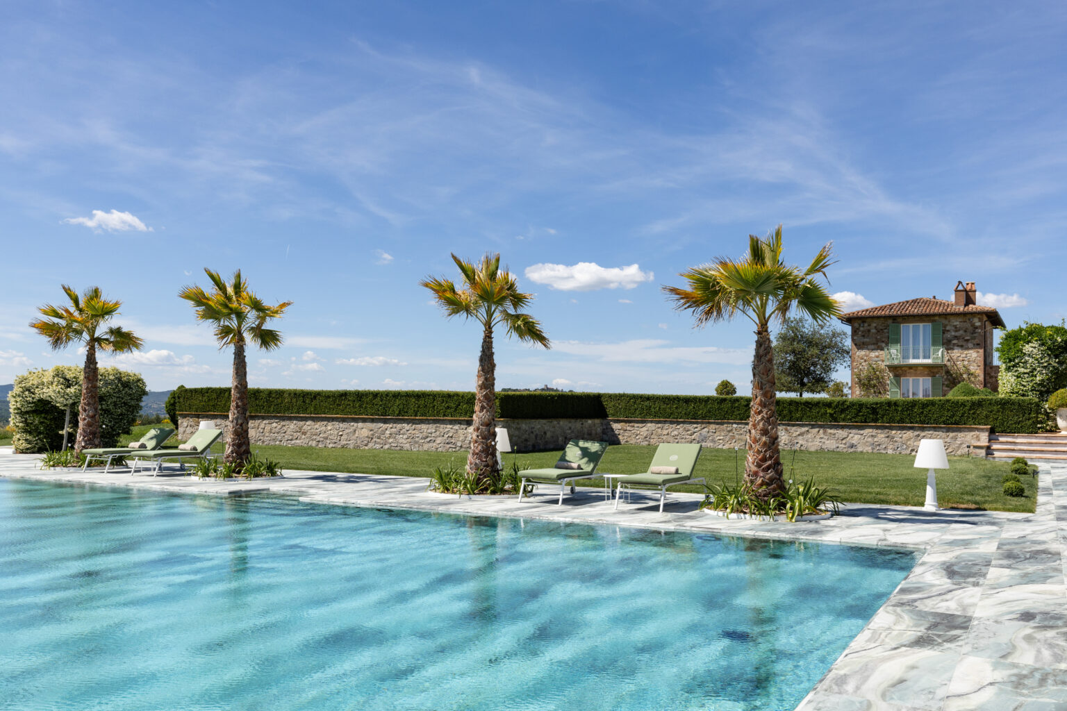 Luxury outdoor swimming pool surrounded by palm trees, sun loungers, and a well-manicured lawn, with a stone house in the background under a clear blue sky. Ideal for relaxation and leisure. | Only on SopranoVillas