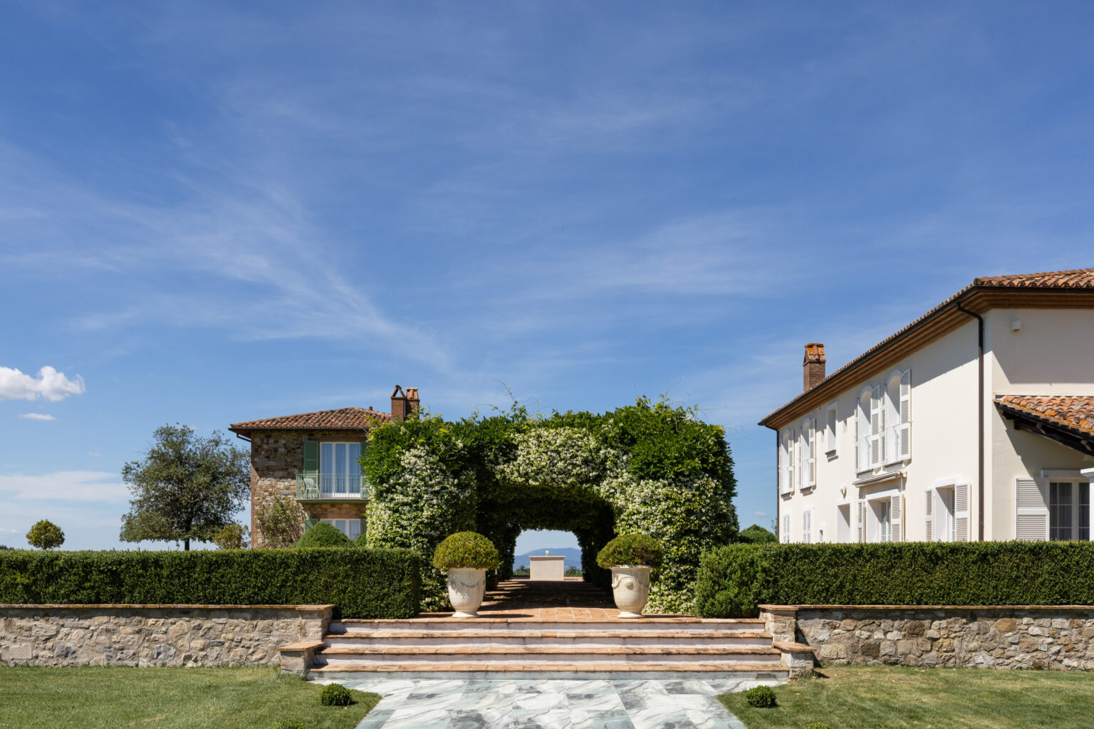Elegant villa entrance with lush greenery and flowering archway, surrounded by manicured hedges and stone pathways, under a clear blue sky. | Only on SopranoVillas